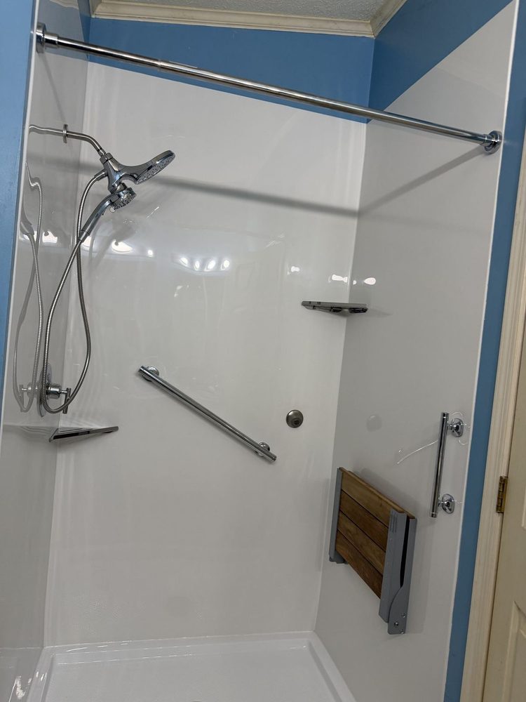 Walk-in shower with white acrylic walls, a handheld shower head, a grab bar and a fold-down wood seat